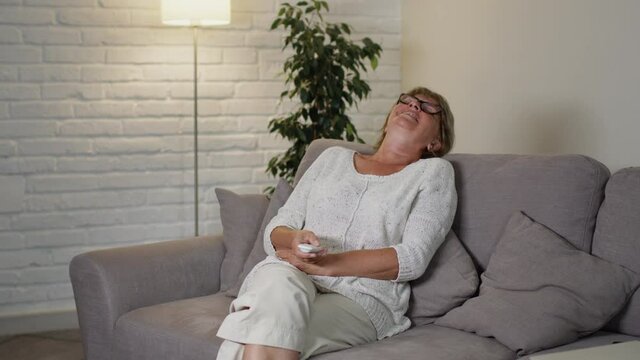 Beautiful Middle Aged Woman Sitting On The Couch In Her Apartment. She Holds The Remote From The TV And Switches Channels. Light In The Room Changes.