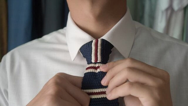 Asian business in formal dress adjusting fashionable knit neck tie then flutter to camera. wardrobe and suit in background.
