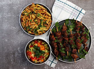 Grilled meat - shish kebabs on a wooden skewers on a round plate served with salad and fried potatoes on a dark background. Top view, flat lay