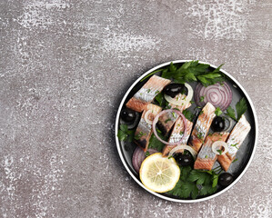 Herring fillet with onions, olives and herbs on a round plate on a dark background. Top view, flat...