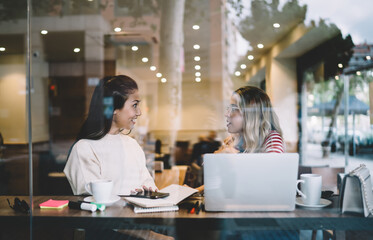 Smiling multiracial female best friends enjoying meeting in cafe talking to each other on coffee break, skilled hipster girls millennials discussing plans for web page publication share blog