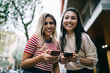Half length portrait of Spanish and Asian millennial girls smiling at camera listening positive music during together pastime, happy tourists in electronic headphones enjoying leisure in city