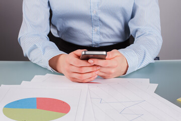 Business woman using smartphone while working with documents. Young business lady surfing internet on mobile phone at her workplace.