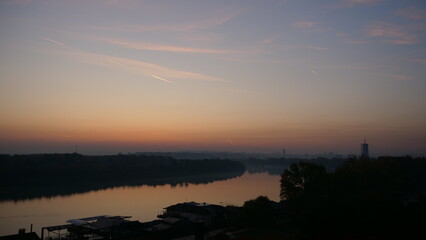 Sunrise Time Lapse on Danube River with Belgrade Serbia Buildings in Background. Blue and Orange Sky with Nature in the Landscape and Trees Reflecting on the Calm Waters of the Danube River.