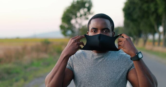 Sweaty African American young handsome sportsman runner looking at camera outdoor at nature and taking on black mask on mouth. Male jogger wearing respiratory protection. Pandemic concept.