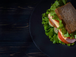 Sandwich with ham, cheese, lettuce, tomatoes, cucumbers on a stone plate on a dark wooden background. Copy space. Top view.