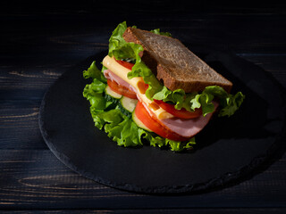 Tasty sandwich with lettuce, tomatoes, cucumbers, ham, cheese on a black stone plate on a dark wooden background. Close up. 