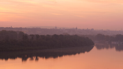 Fototapeta premium Danube River Bank Landscape with Trees Reflecting on Water at Sunrise in Timelapse with Belgrade Serbia City Downtown with Modern and Historical Buildings of Belgrade City Downtown in the Background. 