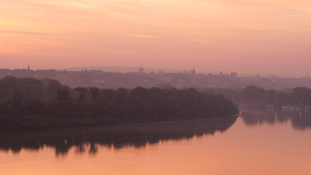 Danube River Bank Landscape with Trees Reflecting on Water at Sunrise in Timelapse with Belgrade Serbia City Downtown with Modern and Historical Buildings of Belgrade City Downtown in the Background. 