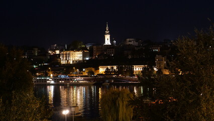 Obraz premium Belgrade Serbia Downtown Night Cityscape Timelapse with Light Reflections on the Sava River Water. The Serbian Orthodox Cathedral Church of St. Michael the Archangel Bell Tower is Visible. 