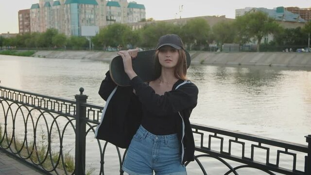 Nice girl putting skateboard on her shoulder while posing on river embankment in happy state of mind.