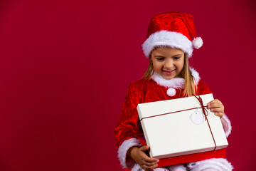 Portrait of a happy young girl with a open gift box isolated over red background, dressed in santa hat and costume