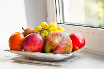 Autumn ripe fruits on the windowsill. Pears, apples and grapes are stacked in a plate near the window. Selective soft focus. Healthy organic food.