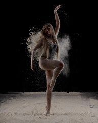 Beautiful sensual fit slim dancer in black body with long blond hair throwing dust, flour, powder on black background.
