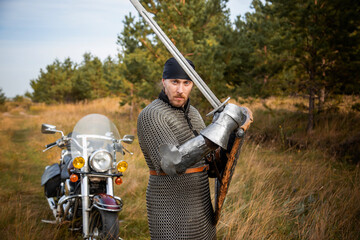 A medieval knight in chainmail with a shield and a sword in his hands stands against the backdrop of a motorcycle and a forest.