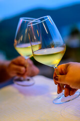Hands with two clinking glasses of cold white wine in mountains cafe at night