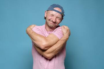 If you are lonely, hug yourself. Cute man in a cap and a pink T-shirt hugs himself and dreams with eyes closed. Blue background.