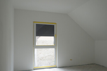 White empty ready-to-paint room with attic ceiling,French window,gray concrete floor.Roller shutter is third down.Design of attic bedroom with sloping roof,panoramic window,Germany.Copy space, mock up
