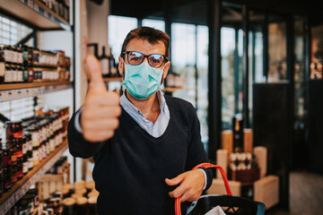 Casually dressed middle age man with face protective mask and gloves buying healthy food and drink in a modern supermarket or grocery store. Pandemic or epidemic lifestyle and consumerism concept.
