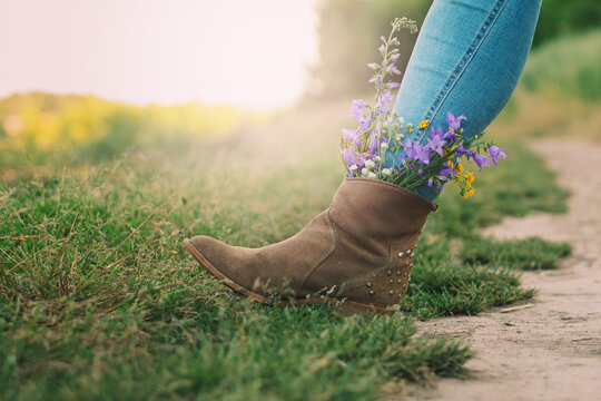 Female legs in jeans high boots with inserted flowers on the road in the field.