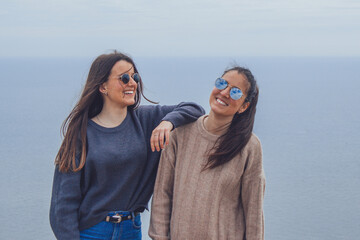 Amigas felices sonríen junto al mar con gafas de sol y ropa abrigada en tarde nublada de invierno