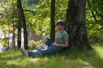 Naklejka na ściany i meble Man sitting under the tree by the lake and using his digital tablet. Guy working outdoors.