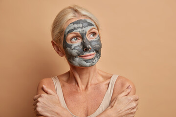 Portrait of thoughtful senior Caucasian woman crosses arms and touches shoulders gently wears homemade facial mask at home concentrated away isolated on beige background. Natural beauty concept