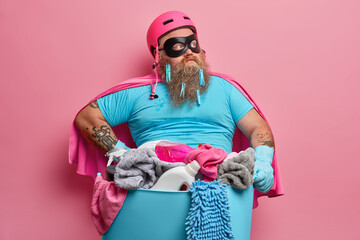 Thoughtful bearded husband in superhero costume has fun and poses near basket full of dirty laundry...