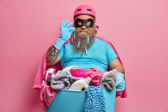 Satisfied busy bearded male housekeeper does laundry at home wears superhero costume wears rubber gloves isolated on pink background poses with basin and cleaning detergents. Housework concept