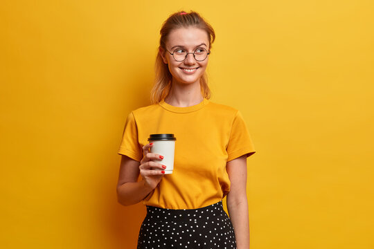 Photo of good looking young European woman holds take away coffee and looks away has cheerful face expression dressed in fashion clothes isolated over yellow studio background. Drinking concept