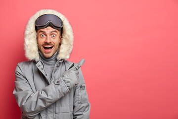 Happy surprised excited man snowboard rider in winter jacket relaxes after skiing has active day of recreation points away on blank space isolated over coral background. Seasonal vacation concept