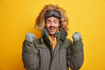 Happy young man rejoices winter came raises clenched fists wears gloves and warm jacket with hood enjoys travel or vacation goes skiing during cold frosty day dressed in wind resistance clothes