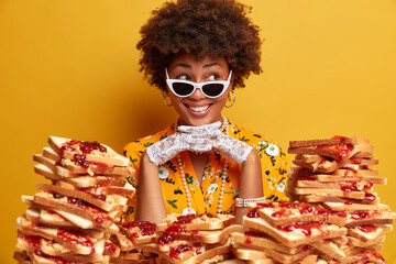 Happy Afro American woman smiles broadly keeps hands under chin and looks with dreamy expression away wears trendy sunglasses poses near pile of bread toasts. Lady spends free time on banquet