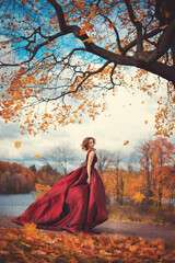 Romantic portrait of a young girl in a long red dress standing in the wind on the background of autumn nature