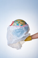 The concept of protecting the world from plastic waste, fighting environmental pollution, a hand in a yellow rubber glove holds a white garbage bag with a globe on a light background.
