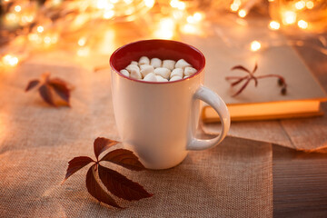 White glass with cocoa and marshmallows on a wooden table with autumn leaves and bokeh in the background. Photos in warm autumn colors.