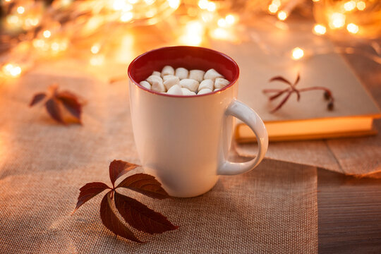 White glass with cocoa and marshmallows on a wooden table with autumn leaves and bokeh in the background. Photos in warm autumn colors.