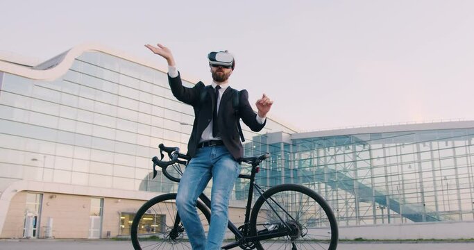 Attractive confident purposeful 30-aged bearded businessman in virtual reality headset standing near his bicycle and working on imaginary screen on the background of modern city building