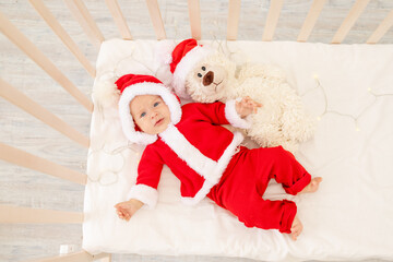 Christmas photo of a baby in a Santa costume lying in a crib at home with a toy in a Santa Claus hat, top view, happy new year