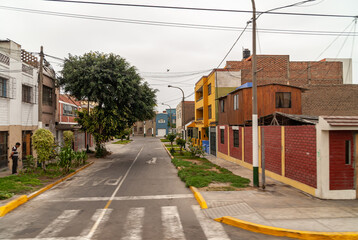 Fototapeta na wymiar Lima, Peru - December 4, 2008: Simple housing on empty street with green and trees on sides in better neighborhood under silver sky. Houses are painted in gray, blue, yellow and red. 