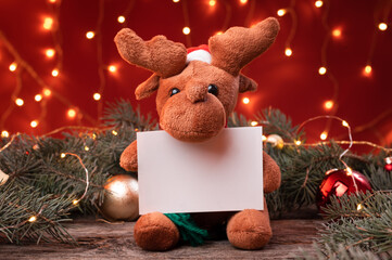 Cute reindeer toy in santa hat holding a white writeable card, sheet of paper in a Christmas...