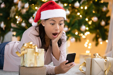 Stunned african American young woman in santa hat lying under Christmas tree shocked by message discount on cellphone, amazed biracial female celebrate new year get surprising text on smartphone