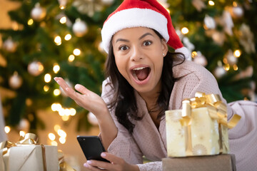 Portrait of shocked biracial woman in Santa hat shocked by Christmas sale or discount message on smartphone, surprised african American female stunned by new year text news on cellphone