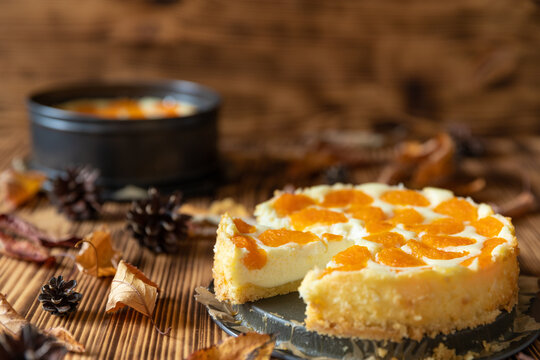 Cheesecake oder K&auml;sekuchen mit fruchtigen orangen Mandarinen auf einem Tisch aus Holz im Herbst mit Laubbl&auml;ttern