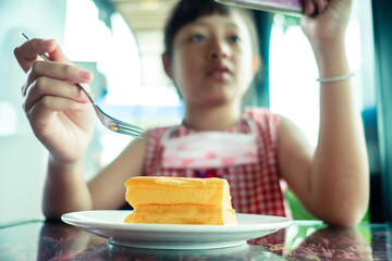Asian little child girl eating chiffon cake with looking smart phone