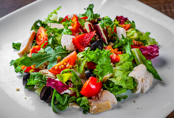 Fresh salad with chicken breast, arugula, black olives,red pepper, lettuce, fresh sald leaves and tomato on a white plate on wooden table background