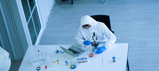 Asian medical scientist wearing protective white jumpsuit and face mask with glasses testing sample with microscope in clinical laboratory. Health care and medical concept. Virus pandemic concept. 