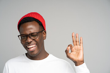 Naklejka premium Cheerful playful young Afro American man wear red hat in good mood smiling broadly, winking, showing okay gesture over studio grey background. Positive Black male in spectacles advertises a product