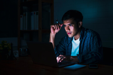 Asian businessman working late night in front using computer laptop smart mobile phone technology, overtime busy schedule focusing hard work at home modern office space feeling tired, sleepy stressful