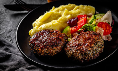 burger with mashed potatoes and vegetables salad on a black plate on Dark grey black slate background